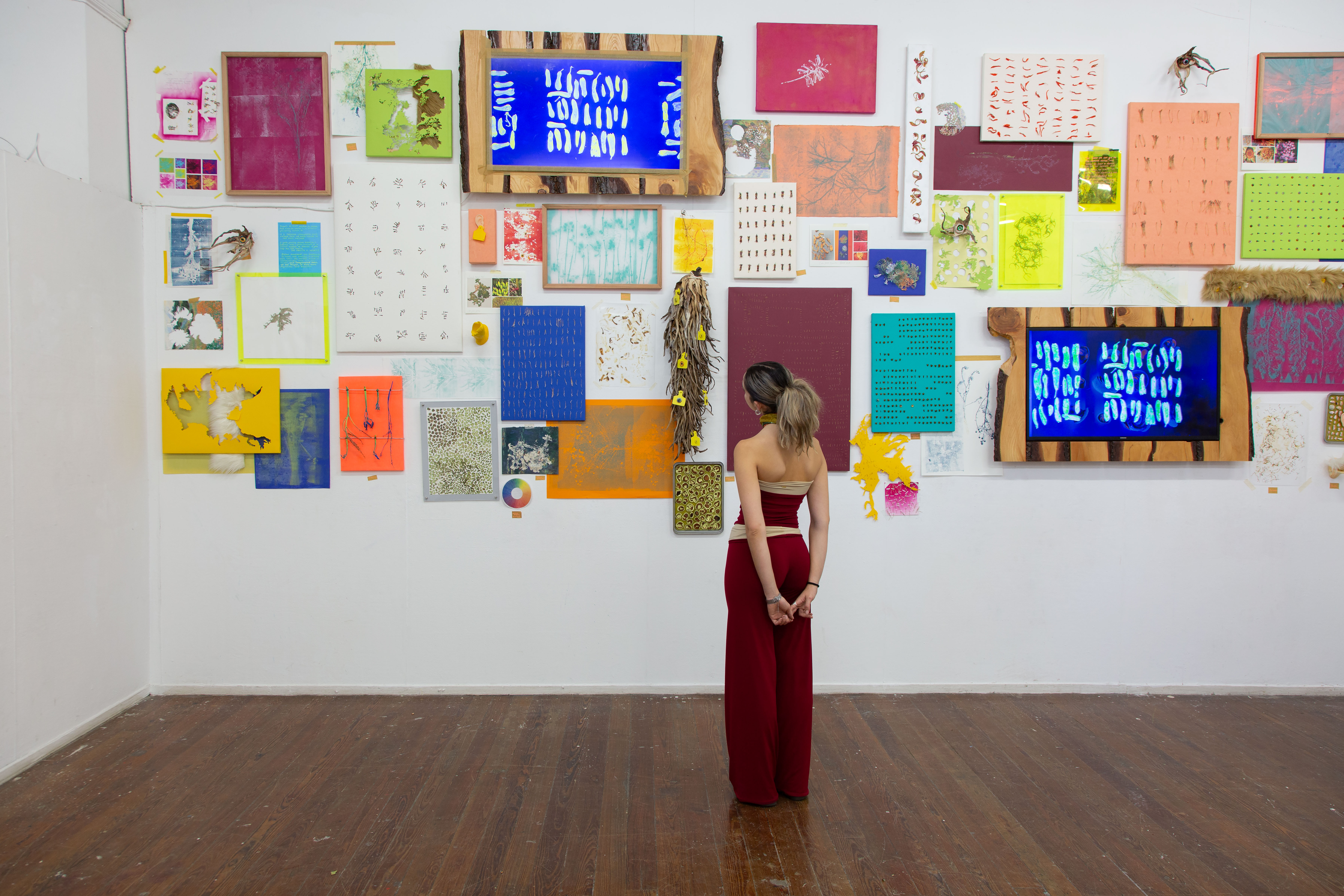 Gabriella Tobler standing in her studio viewing a salon-style installation of colorful artworks including prints, digital screens, and mixed media pieces